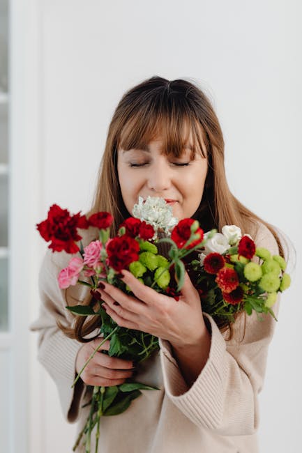 A woman with light brown hair and bangs is holding a vibrant bouquet of fresh flowers close to her face, her eyes gently closed and a content smile. The bouquet features a variety of blooms including