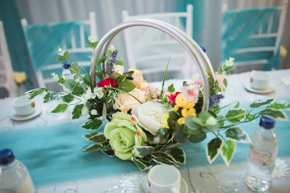 A floral arrangement featuring a mix of soft pink, cream, and white roses, accompanied by purple lavender sprigs, green ivy leaves, and variegated foliage, placed in a circular, metallic basket style 