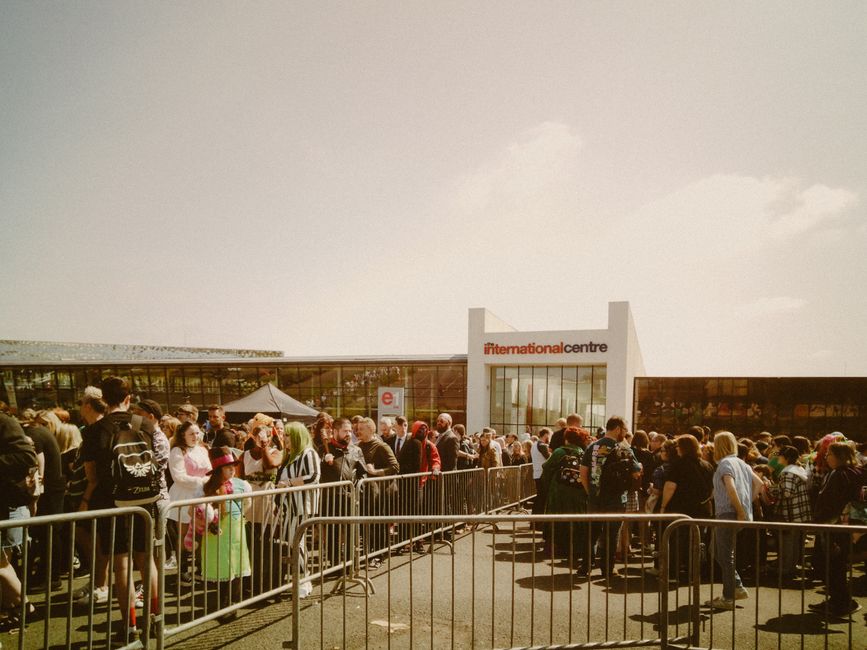 A large crowd of people lined up outside the international centre, standing behind metal barriers on a sunny day with clear skies. The building's modern glass facade and white exterior are visible, wi