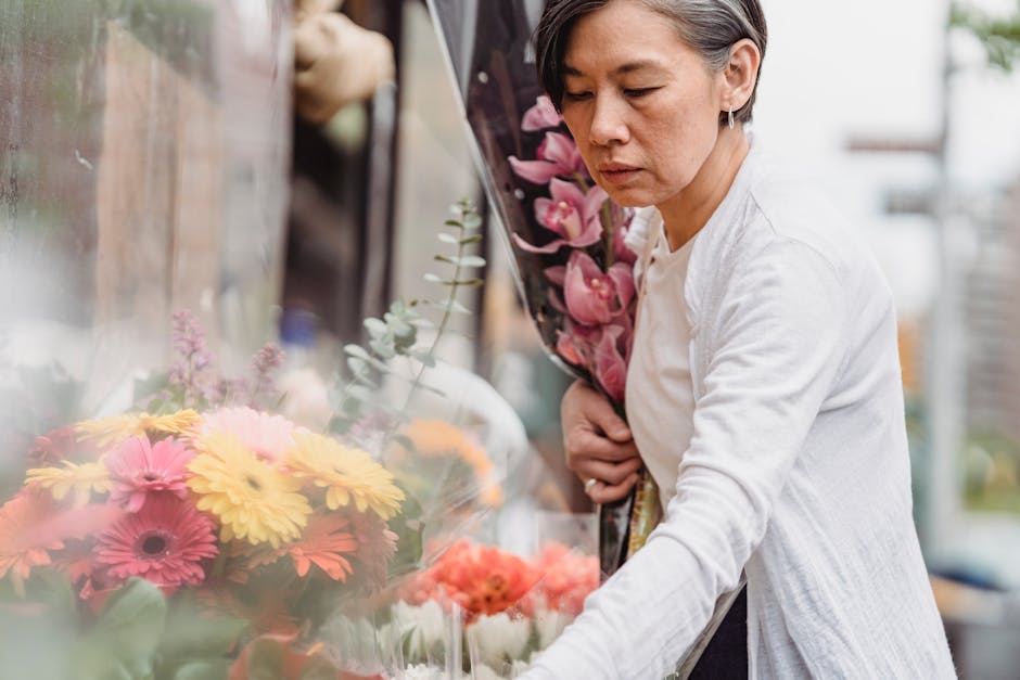 A woman with short gray hair, wearing a light gray cardigan over a white top, selecting a bouquet of fresh flowers at an outdoor flower shop. She is holding a large arrangement of pink orchids wrapped