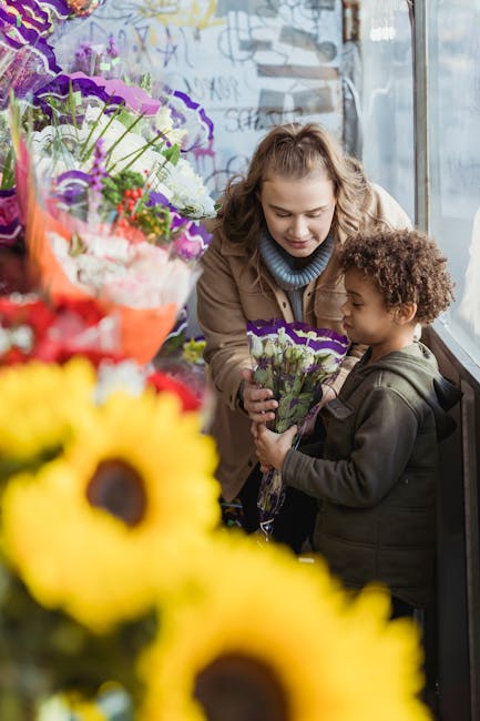A young woman with light brown, wavy hair and a beige coat is handing a small bouquet of purple and white carnations to a curly-haired young boy dressed in a dark hoodie, both smiling softly as they s