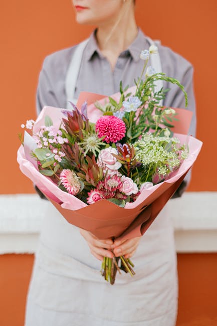 A florist from flowersqueensbury.co.uk holds a vibrant bouquet of fresh flowers arranged with pink, white, and burgundy blooms, including chrysanthemums, roses, and greenery, wrapped in soft pink pape