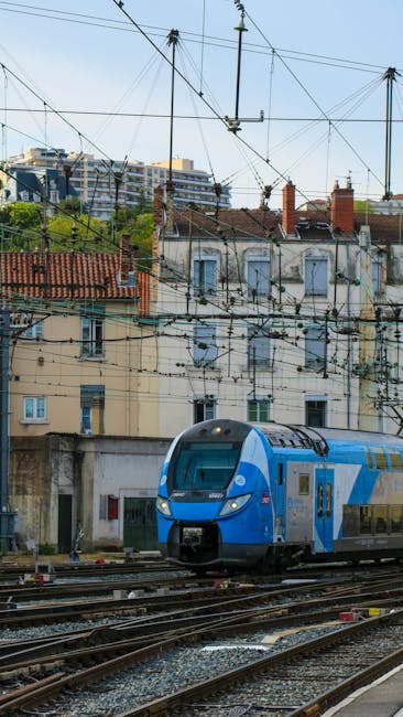 The image shows a modern blue and gray train traveling along railway tracks in an urban area under a network of overhead electric wires. Behind the train, there are old residential buildings with weat