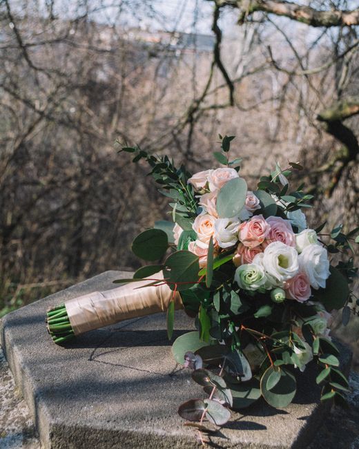 A bouquet of fresh pink and white roses, complemented by white ranunculus and silvery eucalyptus leaves, resting on a stone surface outdoors. The bouquet is wrapped with a light-colored ribbon around 